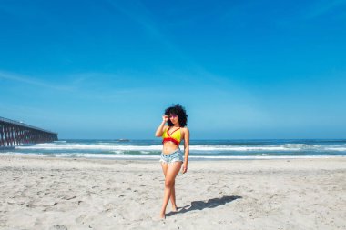 beautiful african american woman on mexican rosarito beach wearing shorts and bikini standing next to pier, latina clipart
