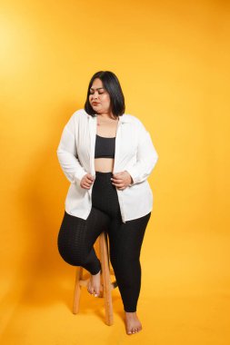 Taburede poz veren, beyaz gömlek ve siyah tayt giyen, kendinden emin, iri yarı bir kadın.