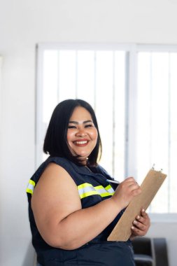 Artı, güvenlik yeleği giyen, gülümseyen ve panoya yazı yazan Latin kadın işçi.