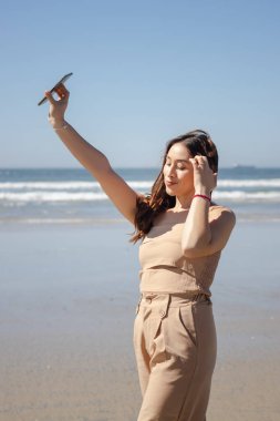 Yaz tatilinin tadını çıkaran genç bir kadın akıllı telefonuyla sahilde selfie çekiyor.