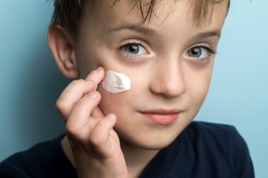 Avrupalı görünümlü bir çocuk kameraya bakar ve kremi yüzüne sürer. mavi arkaplanda dikey fotoğraf