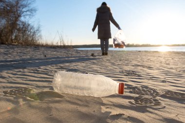 Nehir kıyısında plastik şişe. Çevresel sorunlar ve Dünya Günü. Arka planda çöp torbasıyla gönüllü bir kız.