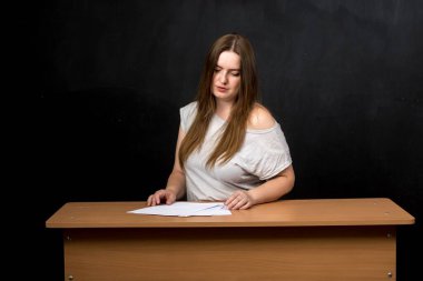 Genç bir bayan öğretmen, arkasında bir karatahta ile bir masada belgelerin üzerine yaslanıyor. Tahtada yazı veya tasarım için yer var. Eğitim, okul veya mesleki temalar için mükemmel