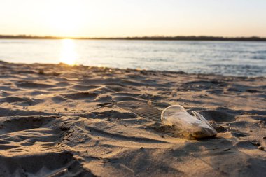 Gün batımında kumlu bir plaja atılan tek kullanımlık plastik bardak çevresel kirliliği simgeliyor. Sıcak arka planda yakalanan resim, nehir veya deniz kıyısındaki atık sorununu vurguluyor