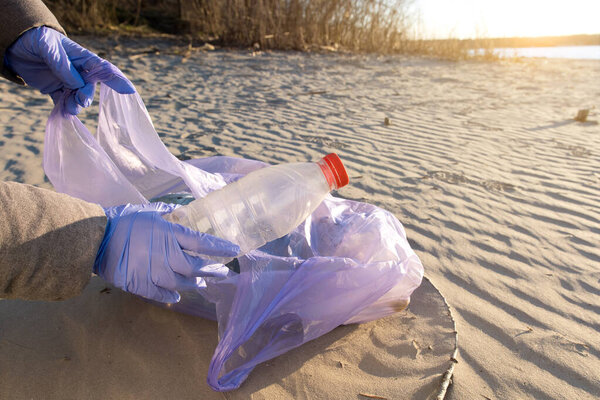 Close-up of a woman's hands in protective gloves putting an empty plastic bottle into a trash bag on a sandy beach. Warm sunlight, with river or sea in the background, highlights beach cleanup action