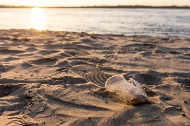 Çöpe atılmış plastik bir bardak gün batımında kumsalda yatıyor. Sıcak güneş ışığıyla aydınlatılan kum yığınının etrafındaki ayak izleri nehir, deniz ve okyanus kıyılarındaki kirlilik sorununu vurguluyor..
