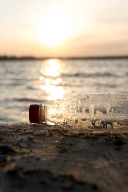 Plastik bir şişe gün batımında kumlu bir plajda uzanıyor ve çevre kirliliği sorununu vurguluyor. Sıcak güneş ışığı, doğanın dinginliğine ve insan etkisine zıt düşer.