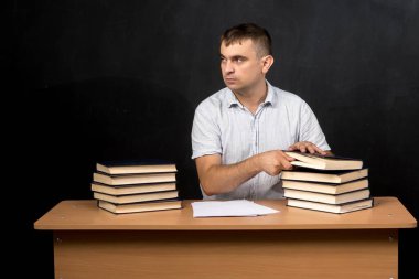 Masada oturan genç bir öğretmen, her iki tarafında kitap yığınları ve önünde kağıtlar. Düşünceli bir şekilde, siyah bir arkaplanla, mesaj ya da tasarım için yer bırakarak. Eğitici içerik için ideal
