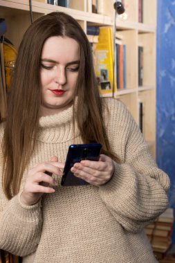 Rahat bir ev ortamında kitaplığa yaslanırken akıllı telefon kullanan genç bir kadın. Sahne, günlük yaşamda modern iletişim ve teknolojiyi vurguluyor. Yaşam tarzı ve teknolojiyle ilgili konular için ideal