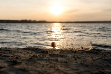 Gün batımında kumlu bir plajda terk edilmiş bir plastik ya da cam şişe, kıyı bölgelerinin kirliliğini simgeliyor. Çevresel hasarı temsil eden çarpıcı bir görüntü ve daha temiz okyanuslar için çağrı