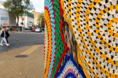 Ağaç gövdesi rengarenk örgü iplikleriyle süslenmiş, şehir yaratıcılığının özünü yakalıyor ve altın sarısı sütunlar çiziyor. Sokak sanatının ve doğanın ahenkli bir gösterisi.
