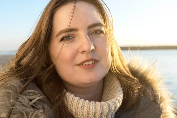 Kışlık ceketli, kumsalda gün batımının yumuşak parıltısıyla gülümseyen bir kadının yakın çekimi. Işık, sakin bir deniz ve arka planda kumlu bir sahil ile onun gevşek yüz ifadesini vurgular.