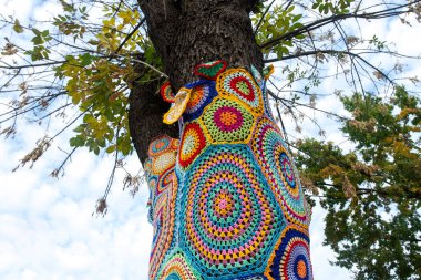 A tree covered in colorful crocheted patterns as part of urban yarn bombing, with vibrant geometric shapes and textures. The creative installation stands out against the natural and urban backdrop clipart