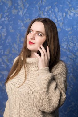 Sıradan bej kazaklı bir kadın cep telefonuyla konuşuyor, yan tarafa bakıyor, mavi arka planda duruyor. İş, kişisel iletişim ya da yaşam tarzı görüntülemesi için uygun
