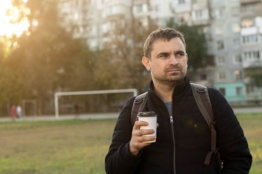 Gün batımında şehir parkında elinde kahve fincanı tutan bir adam. Ceketi ve sırt çantası var. Arka planda bir apartman kompleksi ve bir futbol sahası yer alıyor, şehir yaşam tarzı ve açık hava eğlencelerini temsil ediyor.