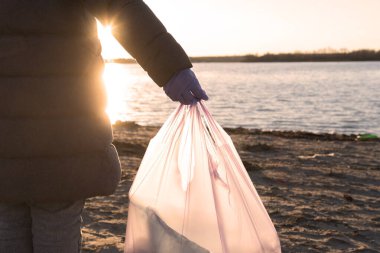 Close-up of a hand holding a trash bag during beach cleanup at sunset. Highlights pollution and environmental care. Perfect for themes of sustainability, ecology, and ocean conservation clipart