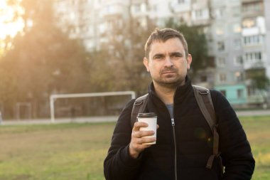 Gün batımında bir parkta elinde kahve fincanı olan bir adam, arkasında bir apartman bloğu olan bir sırt çantasıyla sıcacık giyinmiş. Rahatlama, şehir hayatı ve kahve kültürü konuları için mükemmel.