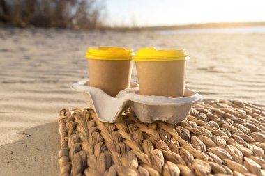 Two takeaway paper cups for coffee or tea on a paper holder, set on a beach at sunset. Ideal for promoting eco-friendly products, featuring blank space for logos or text on the cups clipart