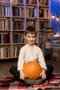 Little boy holding pumpkins in a cozy library setting. The perfect image for illustrating family autumn activities, cozy indoor scenes, and seasonal celebrations. clipart