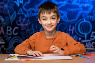 A child in an orange shirt sits at a desk with colored pencils and paper. The background features a chalkboard filled with educational drawings and symbols. Perfect for learning themes.  clipart