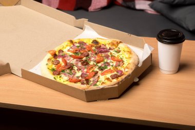 İçinde domatesli, sosisli, kırmızı soğanlı ve otlu pizza olan ahşap bir masanın üzerindeki pizza kutusunu aç. Kutunun yanında siyah kapaklı tek kullanımlık beyaz bir fincan var. Sıradan yemek temaları için mükemmel..