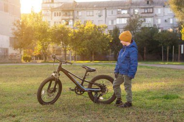 A child stands next to a black mountain bike in a grassy area, wearing a blue jacket, green pants, gloves, and a yellow hat. Trees and apartment buildings are in the background, lit by warm sunlight. clipart