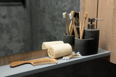 Banyo tezgahında bambu diş fırçaları, sabun makinesi, ahşap tarak ve taş kaplarda pamuk çubukları var. Arka planda ayna ve ahşap bir dolap olan çevre dostu ve doğal bir düzenek.