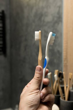 Close-up of a hand holding a wooden toothbrush and a plastic toothbrush. The eco-friendly wooden brush contrasts with the plastic one, emphasizing sustainable choices in personal care. clipart