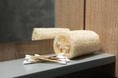 Çevre dostu banyo esasları: gri bir rafta banyo lifi ve bambu pamuklu çubuklar. Arka planda ayna ve ahşap paneller var. Minimalist ve doğal bir görünüm yaratıyor. Sürdürülebilir canlı temaları için ideal.