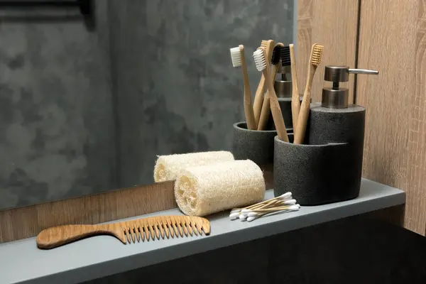 Banyo tezgahında bambu diş fırçaları, sabun makinesi, ahşap tarak ve taş kaplarda pamuk çubukları var. Arka planda ayna ve ahşap bir dolap olan çevre dostu ve doğal bir düzenek.