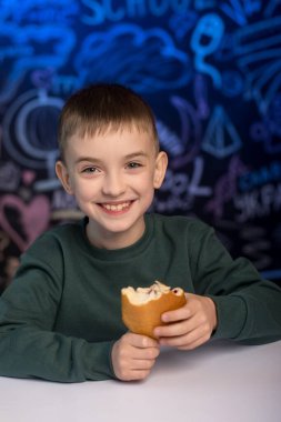 Yeşil kazaklı gülümseyen çocuk okul temalı bir karatahtanın önünde sandviç yiyor. Çocukluk, eğitim ve sağlıklı yemek temaları için mükemmel bir imaj.