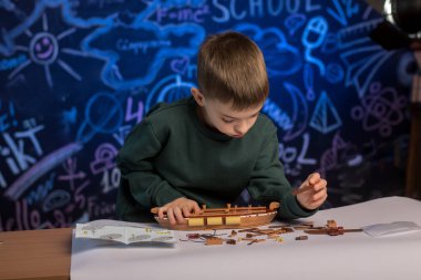 A child in a green sweatshirt building a model ship, immersed in a creative project, symbolizing concentration and practical skills