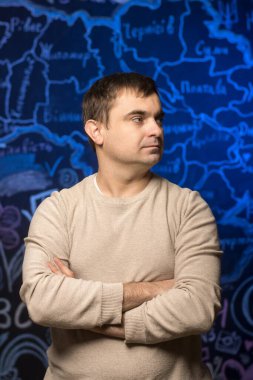 Bej renkli süveter giyen kendine güvenen bir öğretmen mavi haritalı bir arka planda çapraz kollarla duruyor. Eğitim, liderlik ve akademik ilham kavramları için ideal.