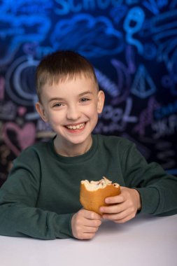 Yeşil kazaklı gülümseyen bir çocuk ısırık almış bir topuz tutuyor, beyaz bir masada renkli bir karatahta arka planına karşı oturuyor. Mutluluk kavramı, çocukluk ve basit mutluluklar.