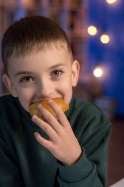 A young boy in a green sweater enjoying a freshly baked bun, sitting indoors against a cozy blue background. The child is smiling softly while holding the pastry close to his mouth, capturing a moment of simple joy clipart