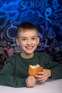 Yeşil kazaklı genç bir çocuk renkli bir okul temalı karatahta arka planıyla sandviç yemekten hoşlanıyor. Eğitim ve okula dönüş kavramları için mükemmel.