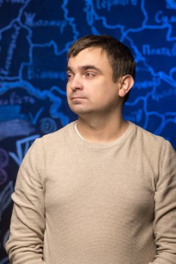 Bej kazaklı bir adam elinde harita olan mavi bir arkaplanda duruyor. Görüntü eğitimsel, profesyonel veya coğrafi temalı içerik için idealdir.