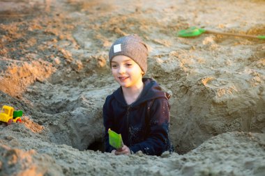 Sahilde kumsalda oynayan bir çocuk elinde yeşil bir kürekle arka planda yeşil bir ağ tutuyor. Sahne gün batımında çekilir, sıcak ve huzurlu bir atmosfer yaratır..