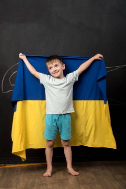 Tahtanın önünde Ukrayna bayrağı taşıyan bir çocuk ulusal gurur ve destek sergiliyor. Mavi ve sarı bayrak belirgin bir şekilde güçlü ve duygusal bir görüntü oluşturuyor..