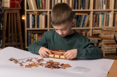 A smiling boy assembling a ship model in a cozy library setting, surrounded by bookshelves. Represents creativity, education, and a love for learning in a peaceful environment clipart