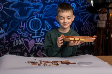 Arka planda çizimlerle kaplı bir karatahtayla gemi modeli yapan gülümseyen çocuk. Okul, eğitim ve DIY hobi temaları için harika.