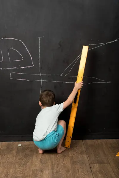 Çocuk elinde büyük bir cetvelle karatahtanın önünde duruyor, okulu ve yaratıcı oyunları vurguluyor..