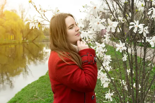 Kırmızı ceketli bir kadın, parktaki dingin bir gölün kenarındaki çiçek açan bir manolya ağacının yanında duruyor ve bahar manzarasının tadını çıkarıyor. Yumuşak güneş ışığı huzurlu atmosferi güçlendirir.