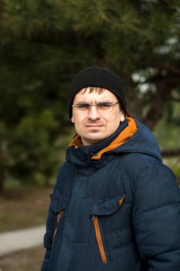 Koyu mavi kışlık ceketli, turuncu aksanlı ve siyah bereli yeşil ağaçların bulanık arka planına karşı duran birinin yakın çekimi. Kış modası ve açık hava etkinlik temaları için ideal