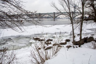 Arka planda çıplak ağaçlar ve karla kaplı bir köprüyle kısmen donmuş bir nehrin yer aldığı sakin bir kış manzarası. Mevsimlik ve doğa temalı projeler için mükemmel.