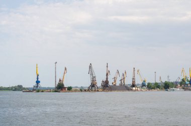 Rıhtım boyunca çeşitli renk ve boylarda büyük vinçleri olan hareketli bir endüstriyel liman, yakınlarda yığınla malzeme ve ön planda parçalı bulutlu bir gökyüzünün altında sakin bir su.