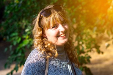 A smiling blonde woman stands in a sunlit park, wearing casual clothes and radiating joy. A perfect capture of a sunny autumn day clipart