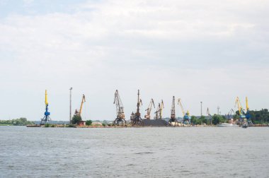Rıhtımda birden fazla vinç bulunan sanayi limanı bulutlu bir gökyüzünün altında deniz lojistiği ve kargo taşımayı gösteriyor. Endüstri, denizcilik ve ulaşım konuları için ideal