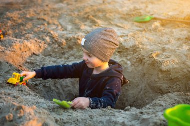 Çocuk kumlu bir plajda oyuncak bir kamyonla dışarıda oyun oynuyordu. Bere ve ceketle sıcacık giyinmişti. Arka plandaki dingin göl ve yeşillik huzur dolu sahneyi tamamlıyor.