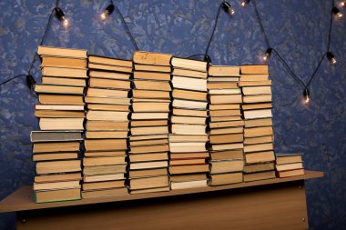 Masada düzgünce düzenlenmiş mavi desenli duvar ve arka planda dekoratif sicim lambaları olan bir yığın eski kitap. Kütüphane, öğrenme ya da rahat okuma alanı kavramları için ideal.